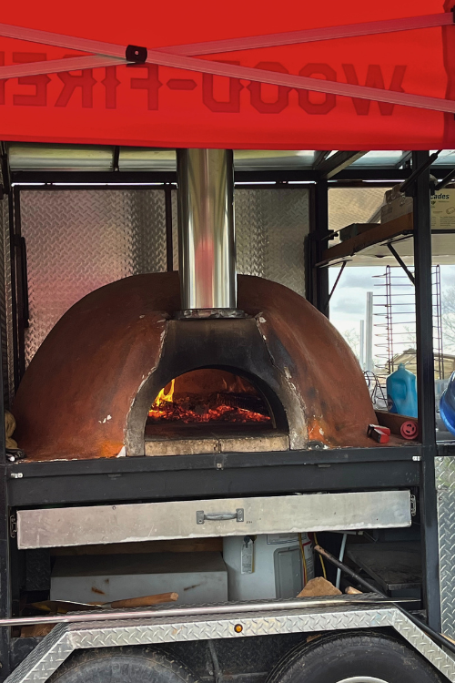 A mobile pizza oven, with red glowing coals, under a red awning that reads 