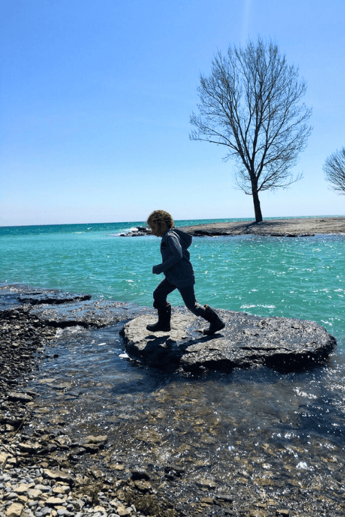 A child jumps across large stones in front of aqua blue water near Pebble Beach, Ontario