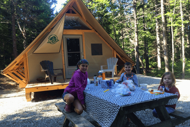 How to Book the Best Campsites in Canada. Children eat breakfast at a picnic table in the woods out front of an oTENTik at the Fundy National Park in New Brunswick Canada.