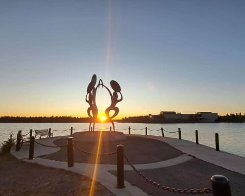 The sunsets over Frame Lake at 10:48pm in Yelloeknife in July. The sun touches the horizon in the distance and shines between a sculpture of people dancing to drums