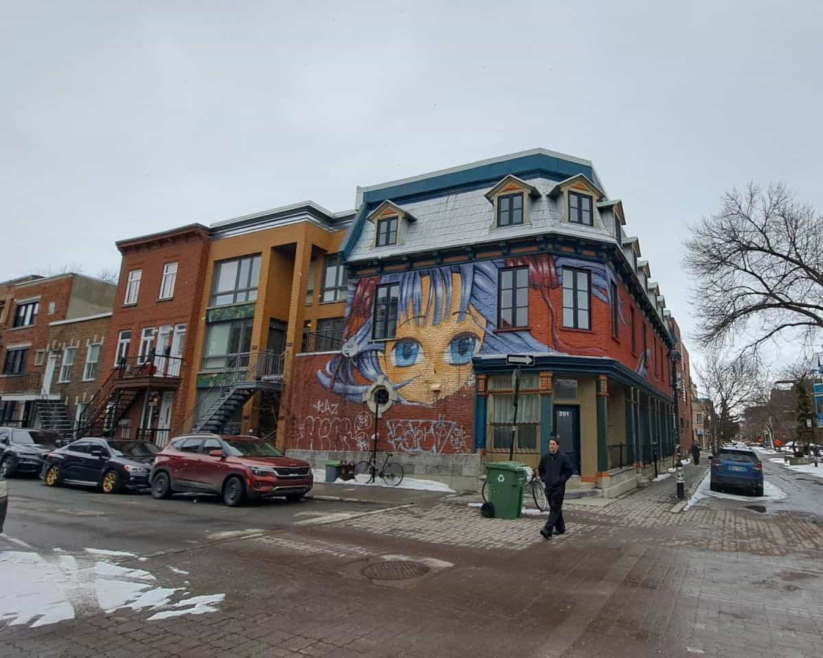 A man crosses a snowy street in the Le Plateau area Montreal, Quebec, Canada. Behind him are some typical Montreal-style row houses with external stairs and balconies. The buildling on the corner has a large mural of an anime style girl's face that wraps around the corner of the building.