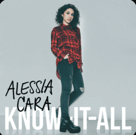 A young woman stands in a empty room, wearing buffalo plaid, standing over the words Know-it-all, Alessia Cara's album cover by the same name.