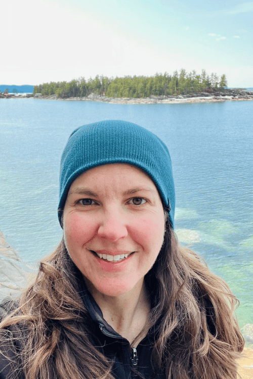 A woman in blue toque smiles in front of the calm blue waters of Agawa Bay, Lake Superior.