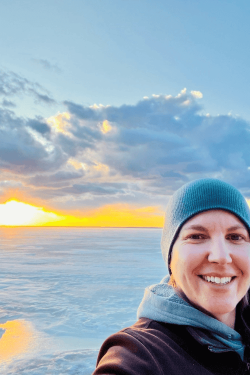 A woman smiles while the sun sets over a lake in the background.