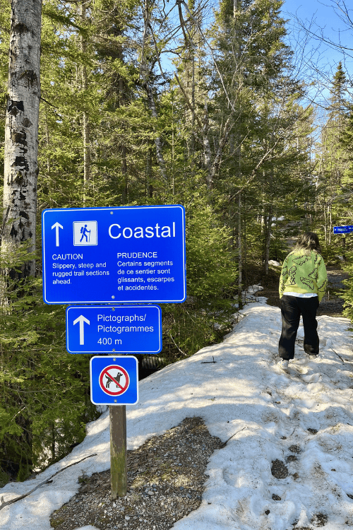 A woman hiking past a Coastal trail sign alerting caution due to slipper, steep and suggest trail sections, pictographs 400m ahead, no dogs. Snow can be seen on the ground.