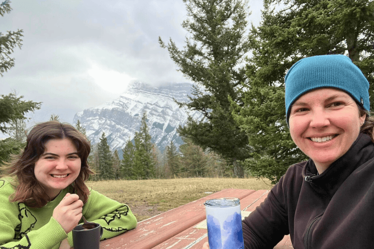 Two woman drink from steaming mugs at a picnic table overlooking snow capped mountain peaks in Banff, Alberta.