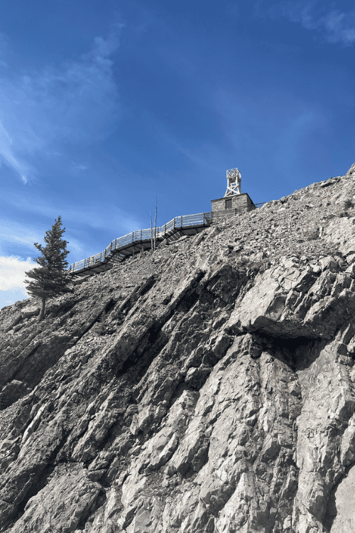 View up the side of a cliff to a wooden structure at the top of a mountain, blue sky day.