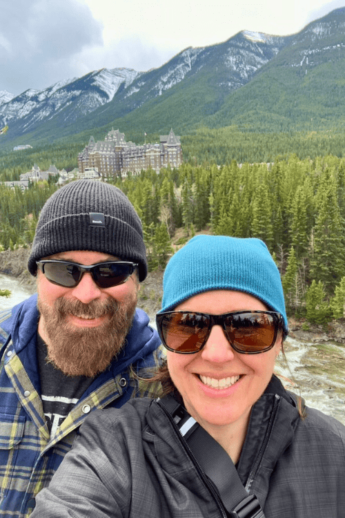 A man and woman smile in front of the Bow River, Banff, Alberta, with Fairmont Banff Springs Hotel in the background