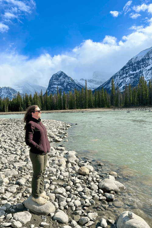 A woman stands near the water's edge in the rocky mountains along the Icefields Parkway, Alberta