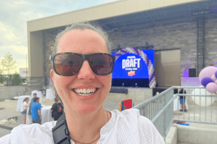 A woman wearing sunglasses smiles in front of the PWHL Draft screen at the outdoor viewing of the PWHL Draft in Ottawa, Ontario