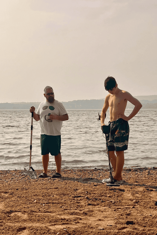 A man and a teen using a metal detector on the shores of a sandy beach on Manitoulin Island, Ontario.