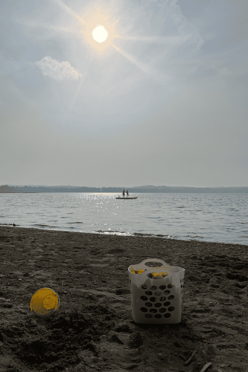 Beach scene including a yellow sand bucket, and white basket filled with sand toys. In the distance two children play on a floating swimming raft and the sun begins its descent.
