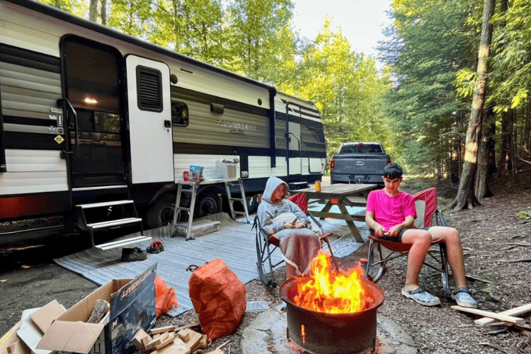 A boy and a girl sit in camp chairs next to a roaring fire by their camper trailer in a wooded campground.