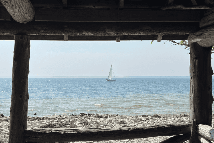 A sailboat glides past the view through an old wood window of a cabin on Flowerpot Island, over the clear blue waters of Georgian Bay, Ontario.