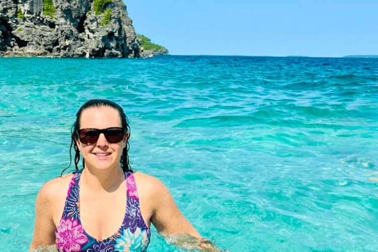 A woman in sunglasses smiles while swimming in the aquamarine waters of Georgian Bay on a blue sky, cloudless day.